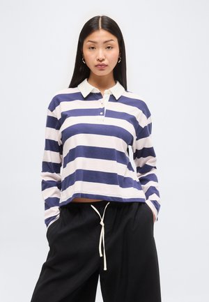 Młoda kobieta z długimi ciemnymi włosami, ubrana w niebiesko-białą pasiastą koszulkę polo z długim rękawem oraz czarne spodnie z troczkami, ręce w kieszeniach.