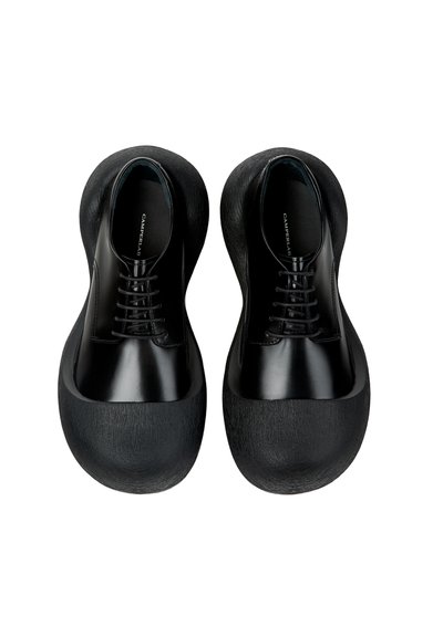 Pari mustia, kiiltäviä nahkakenkiä, joissa on korostetut, pyöristetyt, teksturoidut kumipohjat, kuvattu ylhäältä päin.