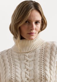 Crème-kleurige kabelgebreide turtleneck trui met een geribbelde kraag en textuurpatronen met verhoogde details in een grove gebreide stof.