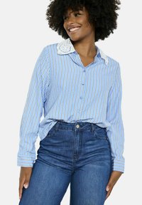 Gai zils garām piedurknēm krekls ar baltu apkakli, vertikālām svītrām un augsto pogu aizdari, apvienots ar augsto jostasvietu tumšiem džinsiem.