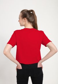 Ung kvinde med brunt hår i en hestehale iført en ensfarvet rød t-shirt og sorte bukser, hænderne i baglommerne, vendt væk fra kameraet.