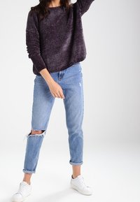 Suéter morado texturizado de corte holgado, combinado con jeans desgastados azul claro y zapatillas blancas. Los jeans presentan desgastes en las rodillas.