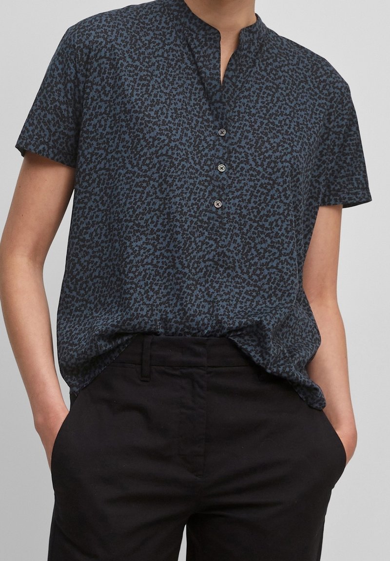 Personne portant un chemisier bleu à manches courtes à motifs avec trois boutons et un pantalon noir, les mains dans les poches.
