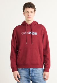 Burgundy hooded sweatshirt with a front pocket, featuring a light blue "Calvin Klein NEW YORK" logo. Soft fabric with ribbed cuffs.