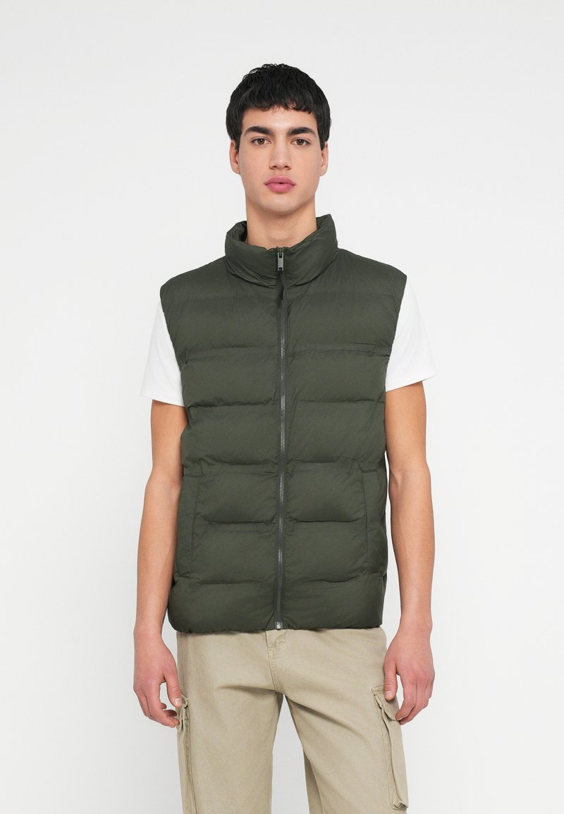 Olivovo zelená prešívaná vesta s vysokým golierom, zapínaním na zips vpredu a prešívanou textúrou, nosená cez biele tričko a v kombinácii so svetlými nohavicami.