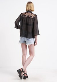 Zwarte doorschijnende blouse met bloemenkantdetails op de rug, 3/4 klokmouwen, gecombineerd met denim shorts en chunky hakken.
