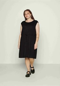 Mulher com cabelo trançado, vestindo um vestido preto sem mangas e sapatos Mary Jane pretos, em pé contra um fundo claro liso.