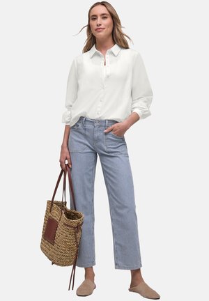 Vrouw draagt een wit overhemd met knopen en blauw gestreepte broek, houdt een geweven tas vast, staat met één hand in de zak.