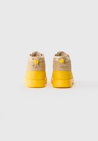 Une paire de bottes en suède beige avec des semelles et des talons en caoutchouc jaune vif, vues de l'arrière sur un fond blanc.