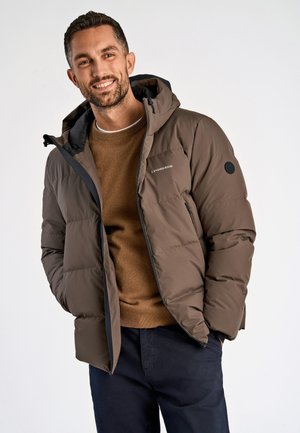 Lächelnder Mann, der eine braune Steppjacke mit Kapuze über einem braunen Pullover und dunklen Hosen trägt, mit den Händen in den Taschen vor einfachem Hintergrund stehend.