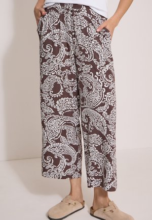 Person iført løse brune bukser med hvide paisley-mønstre og beige slip-on sko, står med hænderne i lommerne mod en neutral baggrund.