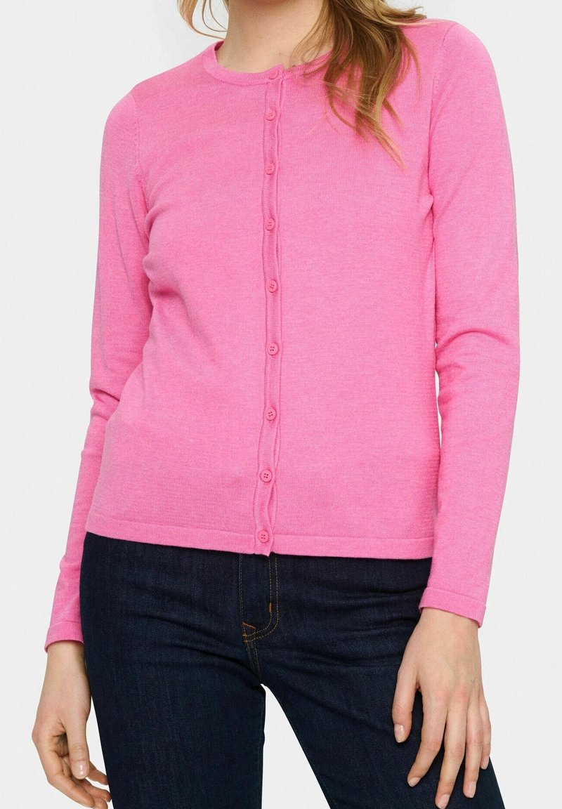 Femme portant un cardigan rose à manches longues boutonné, associé à un jean bleu foncé, debout devant un fond clair uni.
