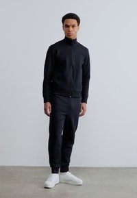 Νέος άνδρας που στέκεται, φοράει μαύρο μπουφάν με φερμουάρ, μαύρο παντελόνι και λευκά αθλητικά παπούτσια, μπροστά σε μια απλή ανοιχτογκρι φόντο.