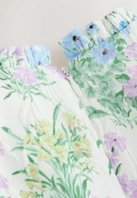 Primo piano di un indumento con motivo floreale con fiori in tonalità pastello giallo, viola e blu, caratterizzato da una zip bianca e bordi del tessuto arricciati.