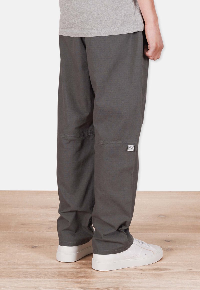 Gray loose-fitting pants with a textured finish, featuring a small logo patch. Worn with white sneakers, displayed against a wooden floor.