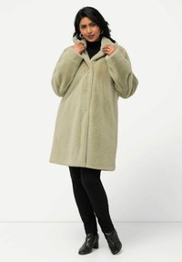 Cappotto in pelliccia sintetica verde chiaro, caratterizzato da una vestibilità rilassata, maniche ampie e un colletto. Abbinato a pantaloni neri e stivaletti con tacco testurizzati.