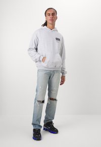 Sweatshirt gris avec poches avant et impression noire, associé à un jean déchiré bleu clair. Des chaussures noires avec des accents bleus complètent la tenue.