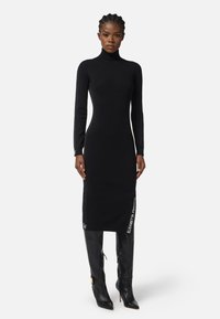 Robe midi noire à manches longues avec un col montant, présentant un tissu texturé et des accents de logo sur le côté. Portée avec des bottes noires hautes.