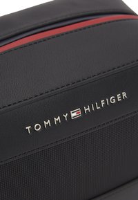 Μαύρη υφασμάτινη τσάντα Tommy Hilfiger με κόκκινη ρίγα και μεταλλικό λογότυπο της μάρκας στο κέντρο του μπροστινού πίνακα.