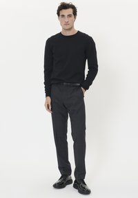 Férfi fekete hosszú ujjú ingben, sötét szürke kockás nadrágban, fekete övvel és fényes fekete cipőben áll, egyik kezét a zsebében tartva.