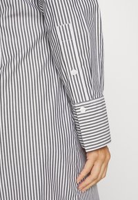 Ingrandimento di una mano appoggiata di lato, che indossa una camicia a maniche lunghe con strisce verticali bianche e nere e bottoni bianchi sui polsini.