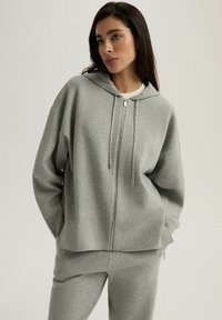 Szara bluza z kapturem na zamek, wykonana z miękkiego materiału. Posiada długie rękawy, sznurki do regulacji oraz luźny krój. Zestawiona z pasującymi szarymi spodniami dresowymi.