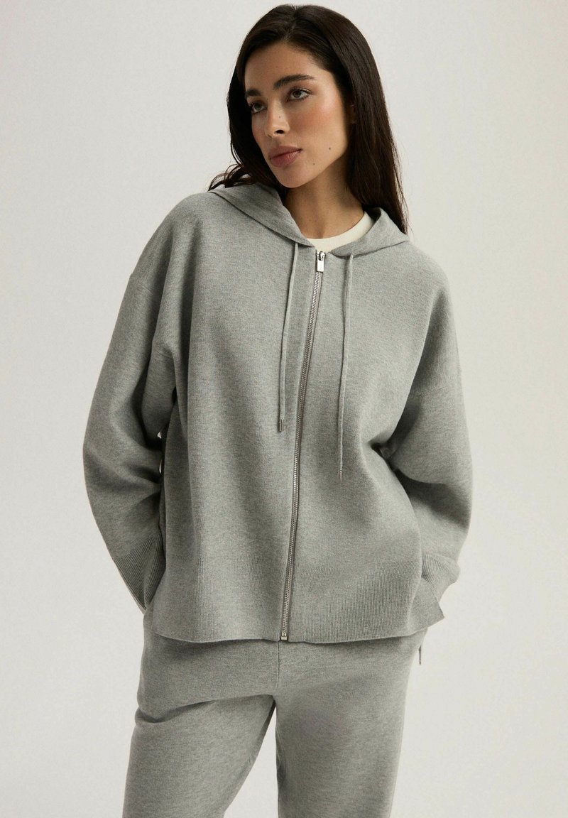 Szara bluza z kapturem na zamek, wykonana z miękkiego materiału. Posiada długie rękawy, sznurki do regulacji oraz luźny krój. Zestawiona z pasującymi szarymi spodniami dresowymi.