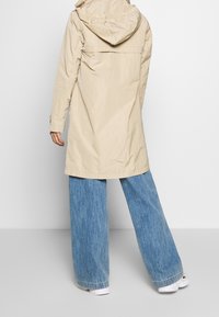 Imperméable long beige avec capuche, en matière légère, doté d'une taille cintrée et d'une coupe droite. Associé à un jean bleu à jambes larges.