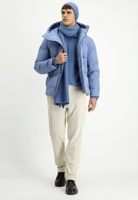Jasnoniebieska pikowana kurtka z kapturem, w połączeniu z niebieskim swetrem, szalikiem i czapką. Beżowe teksturowane spodnie i czarne buty.