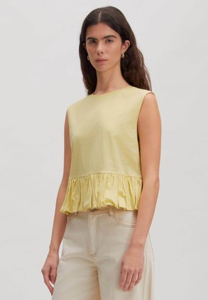 Woman with long dark hair wears sleeveless yellow top with ruffled hem and high-waisted light beige pants standing against gray background.