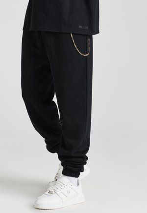 Schwarze Sweatpants in lockerer Passform mit Bündchen an den Knöcheln, ausgestattet mit einem goldenen Kettendetail. Kombiniert mit weißen Turnschuhen und einem schwarzen T-Shirt.