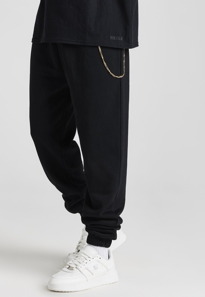 Svart sweatshirtbyxor med avslappnad passform och muddar vid anklarna, med en detalj av en guldkedja. Kombinerat med vita sneakers och en svart t-shirt.