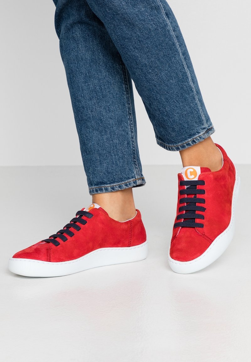 Personne portant des baskets en daim rouge avec des lacets noirs et des semelles blanches, associées à un jean en denim bleu court sur un fond uni.