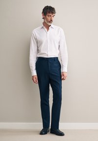 Camicia bianca con bottoni abbinata a pantaloni blu navy a righe sottili e scarpe nere. La vestibilità è sartoriale, con una texture liscia e una silhouette allungata.