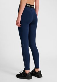 Leggings blu navy con vita nera che presenta il nome del marchio. Il pannello laterale ha un motivo a chevron riflettente. Indossati un paio di scarpe da ginnastica nere.