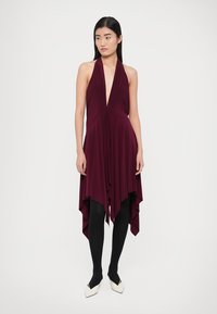 Raudonai vyno spalvos halterio stiliaus suknelė su gilia V formos iškirpte, drapiruota siluetu ir asimetrišku apačios kraštu. Derinama su juodomis pėdkelnėmis ir baltais smailiais batais.