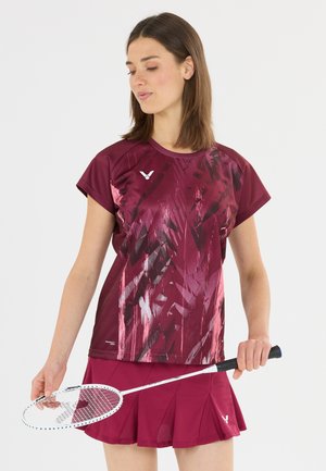 Kvinde iført en bordeaux sportstøj, der holder en hvid badmintonketcher, kigger nedad med en neutral baggrund.