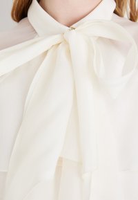 Hvitbluse med myk, transparentt stoff som har høy krage og en stor sløyfe ved halsen, som skaper elegante, flytende linjer.