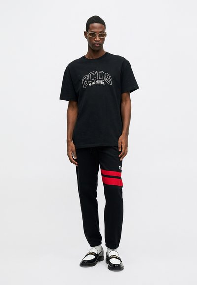 Juoda medvilnė marškinėliai su baltais siuviniais "GCDS" ir logotipu. Juodi džinsai su raudonomis juostelėmis, derinami su baltais kojiniais ir juodais loafers.