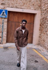 Mężczyzna w brązowej kurtce i jasnych spodniach idący po brukowanej ulicy obok drewnianych drzwi i znaku przejścia dla pieszych.