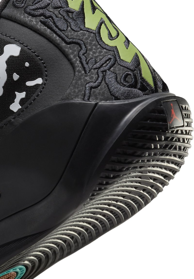 Zapatilla negra con parte superior texturizada que presenta acentos verdes y detalles en blanco. Suela de goma con un patrón de huella distintivo y logo en el talón.
