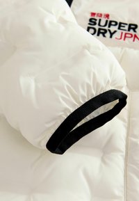 Balta pūkainā jaka piedurkne ar melnu elastīgu aproci, ar kviltu tekstūru un "SUPER DRY JPN" logotipu melnā un sarkanā krāsā.