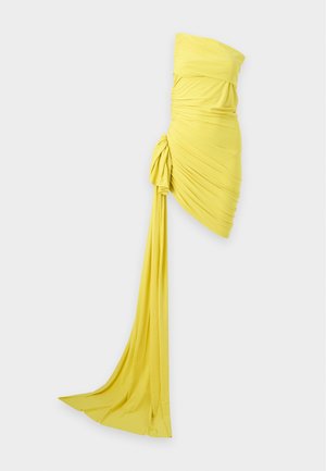 Frau in einem leuchtend gelben One-Shoulder-Raff-Minikleid mit asymmetrischem Saum und langem geknoteten Band, steht in schwarzen Sandalen.