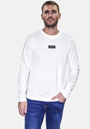 Camiseta blanca de manga larga con cuello redondo y una etiqueta de marca negra en el pecho, combinada con jeans de mezclilla azul oscuro.