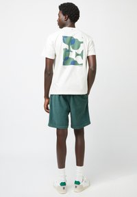 T-shirt bianca con un motivo astratto verde e blu sulla parte posteriore, abbinata a pantaloni corti in corduroy verdi e sneakers bianche con accenti verdi.