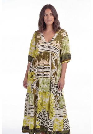 Vrouw die een losse, lange jurk draagt met een V-hals en driekwart mouwen, met een abstract bloemen- en geometrisch patroon in groen, geel en beige tinten.