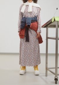 Robe midi à imprimé floral avec manches longues, écharpe blanche et plusieurs vestes superposées nouées à la taille. Baskets jaunes à lacets.