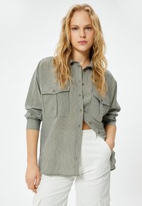 Gestreifte Button-Up-Bluse in Grün und Weiß, mit zwei Brusttaschen, einem lockeren Schnitt und aufgerollten Ärmeln. Kombiniert mit weißen Hosen.