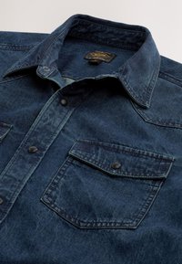 Chemise en denim bleu foncé avec col, boutons-pression et une poche à rabat sur la poitrine, posée à plat sur une surface claire.