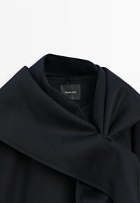 Black coat with a large folded collar, showing a “Massimo Dutti” label inside the neckline on a white background.
